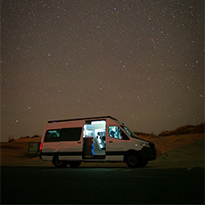 White fourgon aménagé glowing from interior lighting under a starry sky in a remote plage