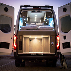 Well-lit fourgon aménagé interior montrant raised bed and illuminated rear gear storage at dusk